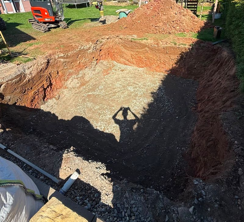 Ausgehobene Baugrube mit Schotterbett und Drainage während der Erdarbeiten, Bagger im Hintergrund
